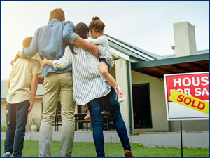man woman and two children hugging in front of home with a sold sign in the yard