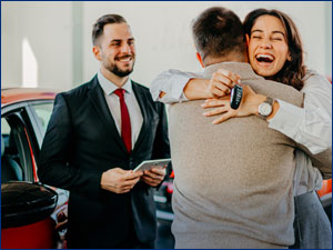 Woman and man hugging after buying a car while car salesman looks on