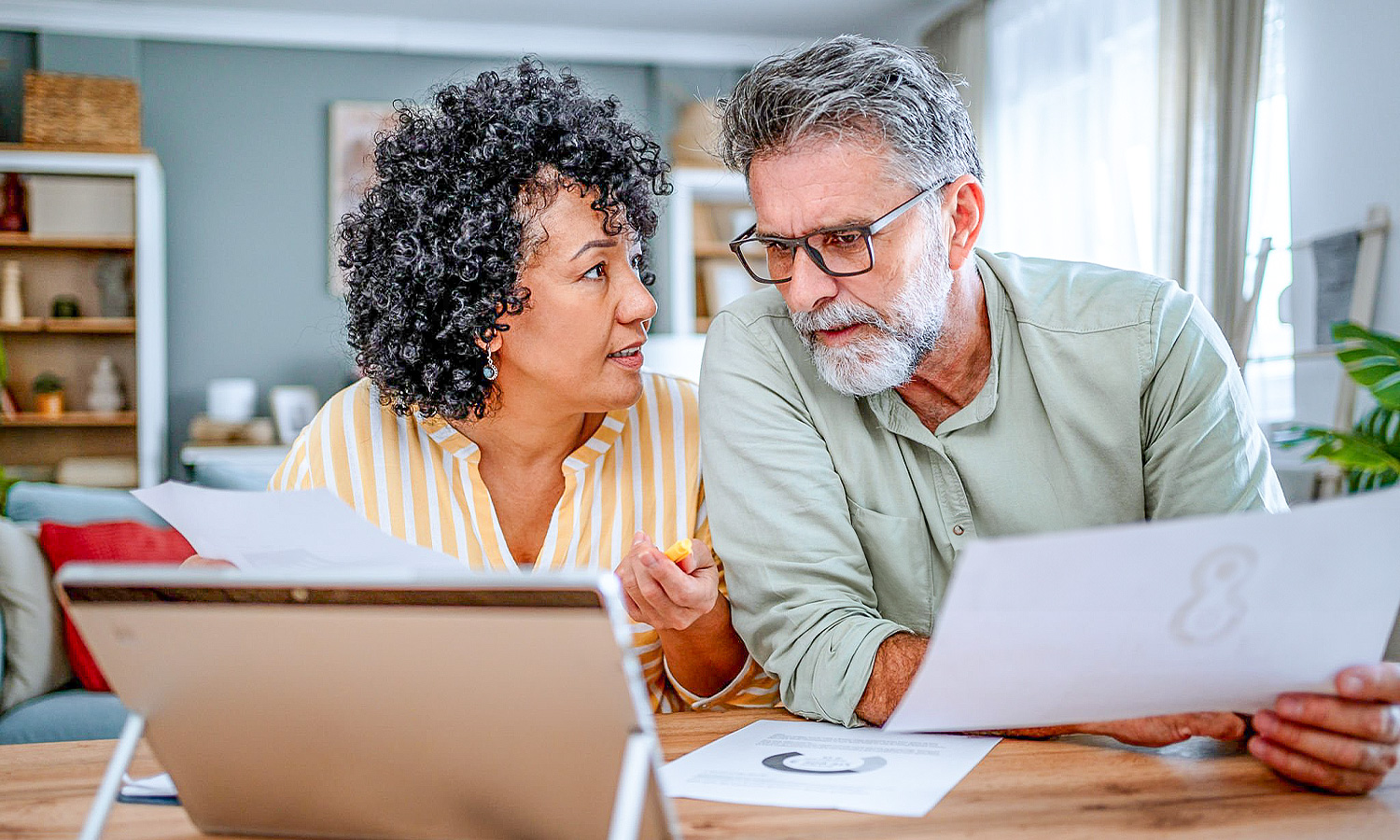woman and man sitting at table curiously looking at tablet and papers with graphs