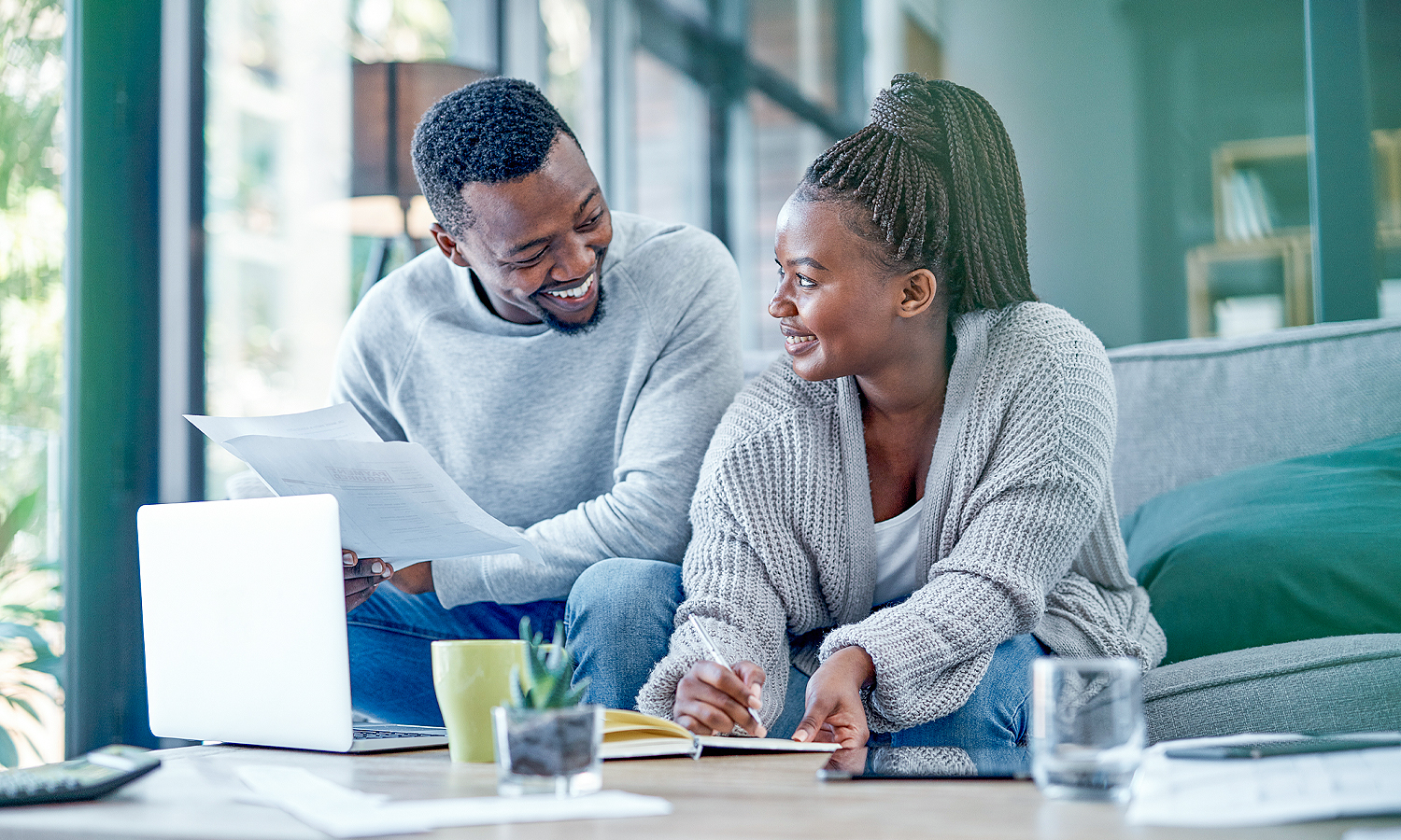 man holding papers and woman writing in notebook while sitting on couch in front of laptop