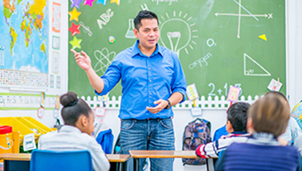 man standing in front of a green chalkboard talking to children sitting in desks